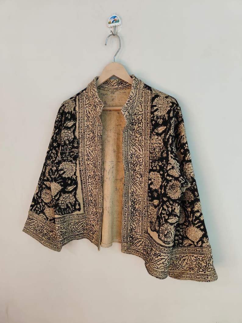 K&ouml;nnte beinhalten: Eine schwarz-beige gemusterte Jacke mit floralem Design h&auml;ngt an einem Holzb&uuml;gel. Die Jacke hat einen hohen Kragen und ist vorne offen, wodurch das beige Innere sichtbar wird. Das florale Muster wiederholt sich auf der gesamten Jacke.