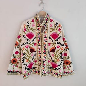 May include: Cream-coloured embroidered jacket with a floral design. The jacket features a high collar and is adorned with pink, orange, and green floral embroidery. The jacket is hanging on a wooden hanger.