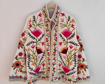 May include: Cream-colored embroidered jacket with a floral design. The jacket features a high collar and is adorned with pink, orange, and green floral embroidery. The jacket is hanging on a wooden hanger.