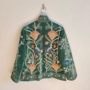 Puede incluir: Chaqueta de terciopelo verde con intrincados bordados florales en tonos melocot&oacute;n, azul y crema. La chaqueta tiene un cuello alto y est&aacute; colgada en una pared blanca.