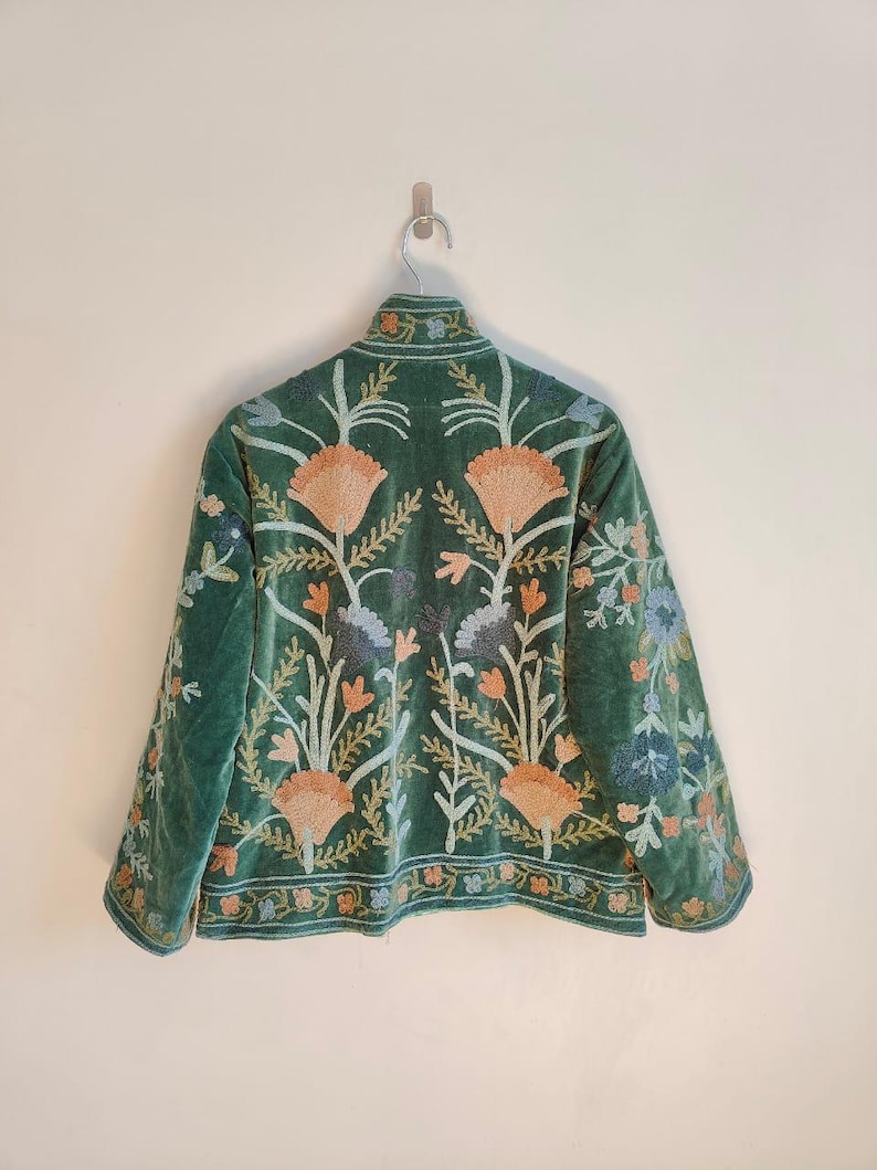 Puede incluir: Chaqueta de terciopelo verde con intrincados bordados florales en tonos melocot&oacute;n, azul y crema. La chaqueta tiene un cuello alto y est&aacute; colgada en una pared blanca.