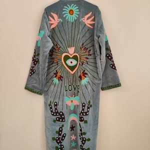Può includere: Lungo abito in velluto blu chiaro con dettagli ricamati colorati. La schiena presenta un cuore, fiori, uccelli e la parola "LOVE". Le maniche e i bordi sono rifiniti in verde. Un capo unico in stile bohémien.