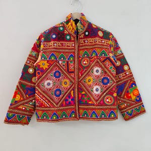 Può includere: Una vivace giacca ricamata in rosso, con intricati motivi geometrici in vari colori. La giacca ha un collo alto e una chiusura con cerniera. Il design incorpora specchi e cuciture dettagliate, creando un aspetto festoso e ornato.