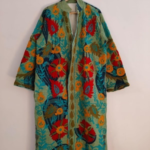 May include: A long, open-front coat with detailed floral embroidery. The coat has a light green base, adorned with red, orange, and teal floral patterns. The embroidery is intricate, giving a textured look. The coat is suspended from a wooden hanger.
