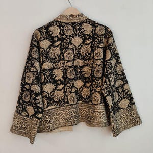 K&ouml;nnte beinhalten: Eine schwarze und beige Jacke mit floralem Muster, die an einem Holzb&uuml;gel h&auml;ngt. Die Jacke hat ein sich wiederholendes Blumenmuster und eine dekorative Bord&uuml;re am unteren Rand. Die Jacke ist aus einem weichen, leichten Stoff gefertigt.