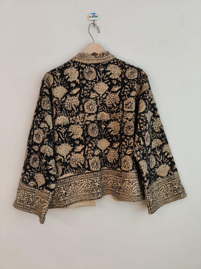 K&ouml;nnte beinhalten: Eine schwarze und beige Jacke mit floralem Muster, die an einem Holzb&uuml;gel h&auml;ngt. Die Jacke hat ein sich wiederholendes Blumenmuster und eine dekorative Bord&uuml;re am unteren Rand. Die Jacke ist aus einem weichen, leichten Stoff gefertigt.