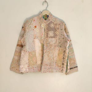 Può includere: Giacca patchwork beige con collo alla coreana, appesa a un'appendiabiti. La giacca presenta vari motivi floreali e geometrici ricamati in tonalità beige, rosa e verde. Le maniche sono lunghe e la giacca è abbottonata.