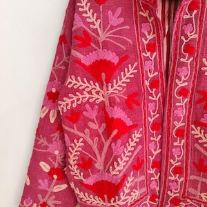 May include: A magenta jacket with intricate floral embroidery in shades of red, pink, and cream. The jacket features a mandarin collar and long sleeves. The floral design is concentrated on the front, sleeves, and collar, creating a detailed and artistic look.