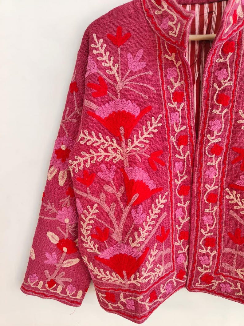 May include: A magenta jacket with intricate floral embroidery in shades of red, pink, and cream. The jacket features a mandarin collar and long sleeves. The floral design is concentrated on the front, sleeves, and collar, creating a detailed and artistic look.