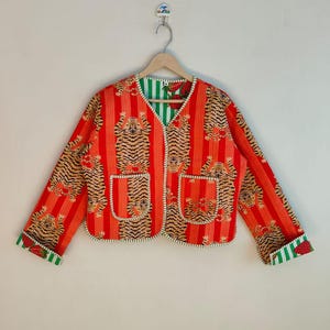 Könnte beinhalten: Eine rot-orangefarbene Jacke mit Tigerstreifenmuster und zwei Vordertaschen. Die Jacke hat einen V-Ausschnitt und ist mit grün-weiß gestreiftem Stoff gefüttert. Die Manschetten weisen ebenfalls grün-weiße Streifen auf. Die Jacke hängt an einem Holzbügel.