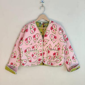 May include: A pink quilted jacket featuring a fig pattern. The jacket has a V-neck, two front pockets, and green trim. The lining is green with a leaf pattern. The jacket is hanging on a wooden hanger.