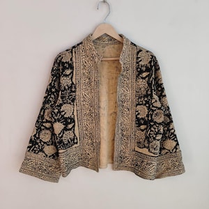 Pode incluir: Uma jaqueta preta e bege com um padrão floral e geométrico. A jaqueta é aberta na frente e tem mangas compridas. A jaqueta está pendurada em um cabide de madeira.
