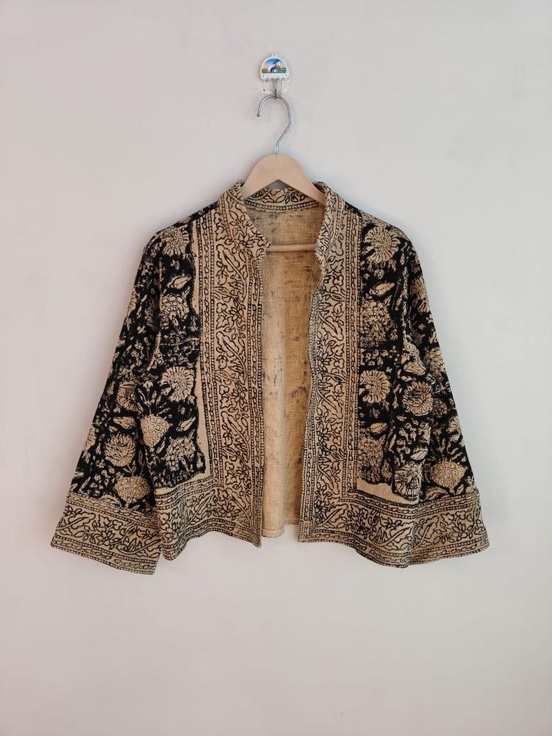 K&ouml;nnte beinhalten: Eine schwarz-beige Jacke mit floralem und geometrischem Muster. Die Jacke ist vorne offen und hat lange &Auml;rmel. Die Jacke h&auml;ngt an einem Holzb&uuml;gel.