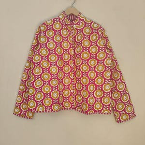 May include: A patterned shirt with a floral design. The shirt has a repeating pattern of yellow and white flowers on a pink background. The long-sleeved shirt is hanging on a wooden hanger.