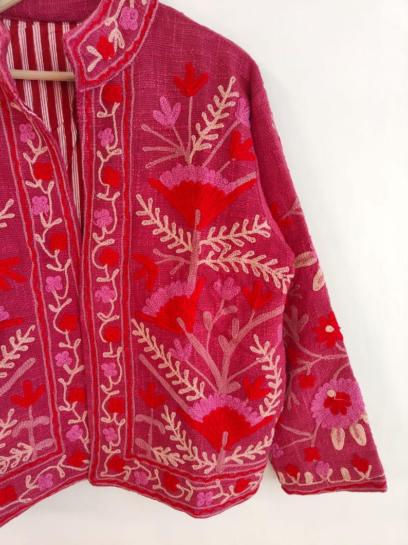 May include: A magenta jacket with floral embroidery in shades of red and pink. The jacket features a mandarin collar and long sleeves. The interior lining is striped. The embroidery includes flowers and leaf patterns.
