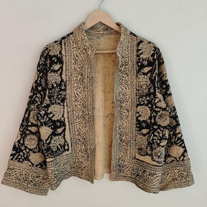 K&ouml;nnte beinhalten: Eine schwarz-beige gemusterte Jacke mit floralem Design. Die Jacke ist vorne offen und hat lange &Auml;rmel. Sie h&auml;ngt an einem Holzb&uuml;gel vor einem wei&szlig;en Hintergrund.