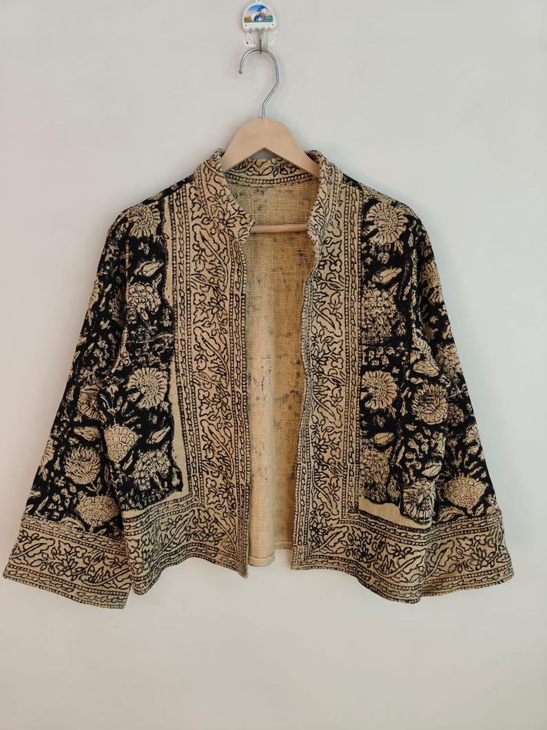 K&ouml;nnte beinhalten: Eine schwarz-beige gemusterte Jacke mit floralem Design. Die Jacke ist vorne offen und hat lange &Auml;rmel. Sie h&auml;ngt an einem Holzb&uuml;gel vor einem wei&szlig;en Hintergrund.