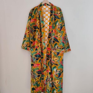 Może przedstawiać: Długi, musztardowy szlafrok z żywym wzorem kwiatowym i liściastym w odcieniach różu, błękitu i zieleni. Szlafrok ma białe wykończenia i podszewkę w kropki. Wisi na drewnianym wieszaku.