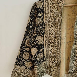 K&ouml;nnte beinhalten: Beige und schwarze Jacke mit floralem und geometrischem Muster. Die Jacke hat einen Stehkragen und Dreiviertel&auml;rmel. Der Stoff scheint ein gewebtes Material zu sein.