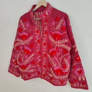 May include: A magenta jacket with floral embroidery in shades of red and pink. The jacket features a mandarin collar and long sleeves. The interior lining has vertical stripes. The jacket is hanging on a wooden hanger.
