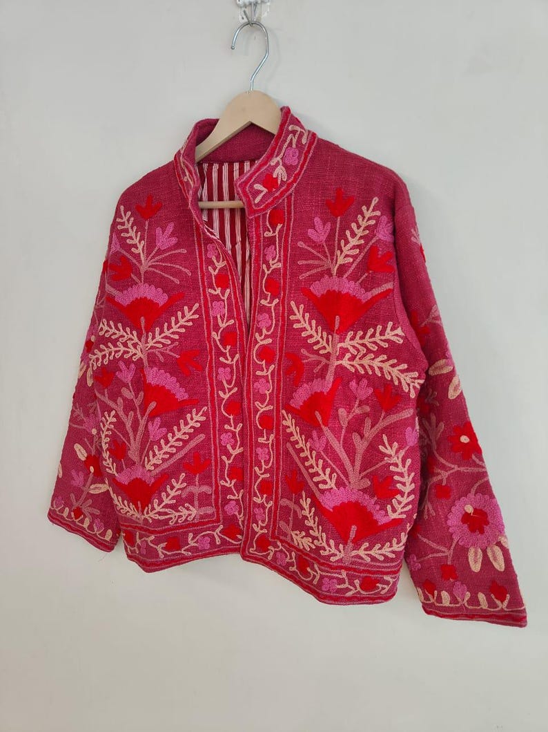 May include: A magenta jacket with floral embroidery in shades of red and pink. The jacket features a mandarin collar and long sleeves. The interior lining has vertical stripes. The jacket is hanging on a wooden hanger.