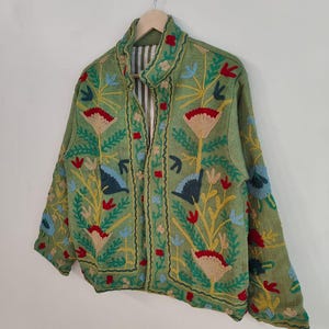 May include: A green jacket with colorful floral embroidery. The jacket features a collared neckline and a striped lining. The embroidery includes red, yellow, blue, and beige flowers and leaves. The jacket is hanging on a wooden hanger.