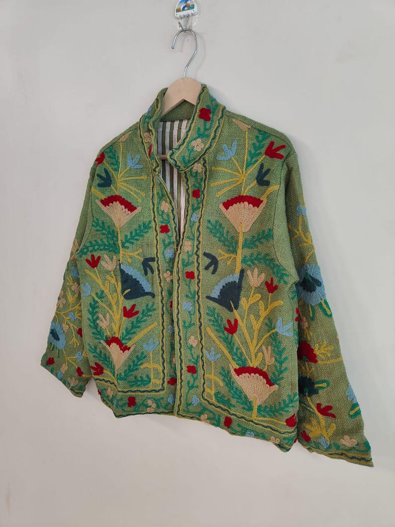 May include: A green jacket with colorful floral embroidery. The jacket features a collared neckline and a striped lining. The embroidery includes red, yellow, blue, and beige flowers and leaves. The jacket is hanging on a wooden hanger.