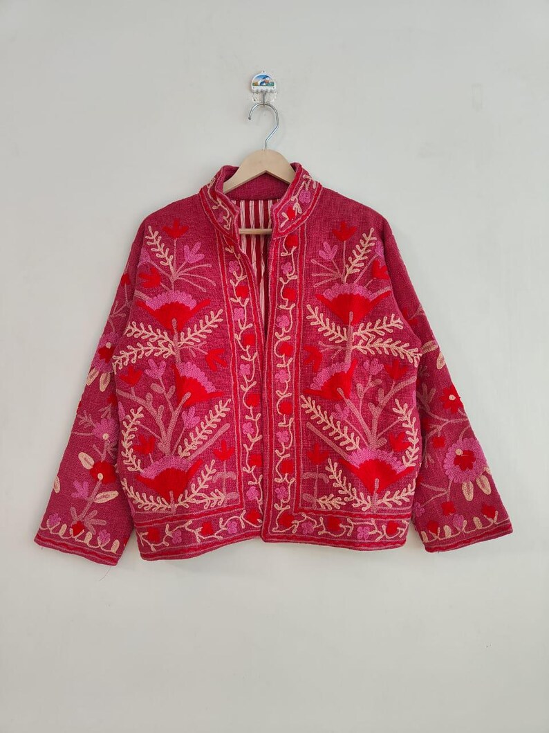 May include: A magenta jacket with intricate floral embroidery in shades of red and pink. The jacket features a mandarin collar and long sleeves. The embroidery includes flowers and leaf patterns, creating a detailed design.