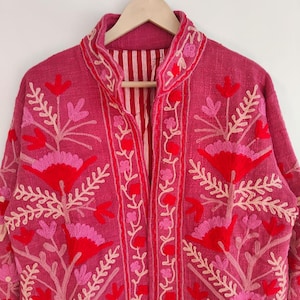 May include: A magenta jacket with intricate floral embroidery in shades of red and pink. The jacket features a mandarin collar and is hanging on a wooden hanger. The interior lining has a striped pattern.