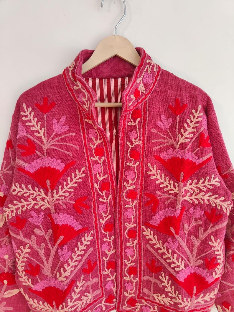 May include: A magenta jacket with intricate floral embroidery in shades of red and pink. The jacket features a mandarin collar and is hanging on a wooden hanger. The interior lining has a striped pattern.
