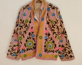 May include: A tan jacket with colorful floral embroidery. The jacket features a high collar and long sleeves. The embroidery includes pink, blue, green, and yellow flowers with brown stems and leaves. The jacket is hanging on a hanger.