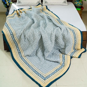 May include: A patterned quilt in shades of blue and cream, draped over a bed. The quilt features a repeating floral design and a contrasting border in a darker blue. The edges are trimmed with a cream-colored fabric. The quilt is approximately 203 cm by 229 cm.
