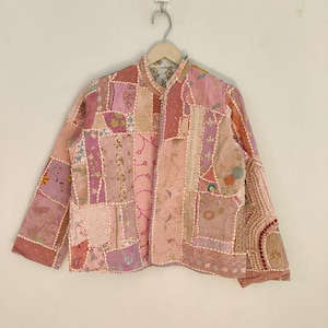 Può includere: Una giacca patchwork nei toni del rosa, lavanda e beige è appesa a un gancio. La giacca presenta vari modelli e trame di tessuto, tra cui stampe floreali e dettagli ricamati. La giacca ha maniche lunghe e un colletto alla coreana.