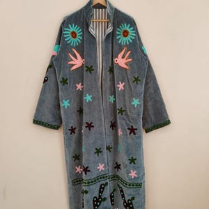 May include: A long, open-front robe in a muted blue-grey colour, decorated with embroidered flowers, birds, and stars in teal, pink, and brown. The robe has a striped lining and green trim on the sleeves and hem, creating a bohemian aesthetic.