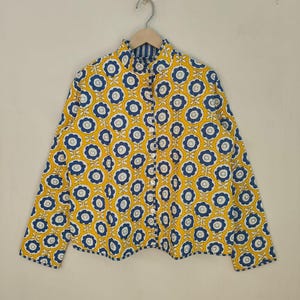 May include: A yellow shirt with a blue and white floral pattern. The shirt has long sleeves and a mandarin collar. The floral design is repeated across the fabric. The shirt is hanging on a wooden hanger.