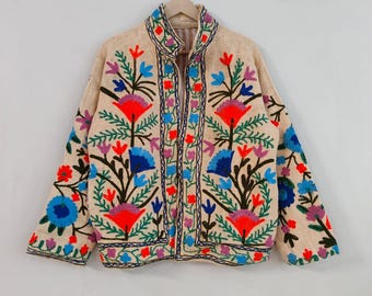 May include: Beige jacket with colorful floral embroidery. The jacket features a high collar and a zipper closure. The embroidery includes red, blue, purple, and green flowers and leaves. The jacket is hanging on a wooden hanger.