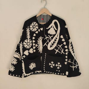 Puede incluir: Chaqueta negra con bordados florales blancos. La chaqueta tiene dos bolsillos delanteros y un forro a rayas. El bordado incluye varios diseños florales y abstractos. La chaqueta está colgada de una percha de madera.