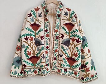 May include: Cream-colored embroidered jacket with a floral design. The jacket features a stand-up collar and is adorned with colorful embroidered flowers, leaves, and stems in shades of red, green, and blue. The jacket is hanging on a wooden hanger.