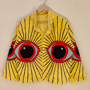 May include: A yellow jacket with a unique eye design. The jacket features large, stylized eyes in red, black, and light blue, with radiating black lines. The jacket is hanging on a wooden hanger.