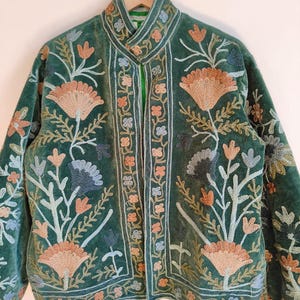 Puede incluir: Chaqueta de terciopelo verde con intrincados bordados florales en tonos melocot&oacute;n, azul y crema. La chaqueta presenta un cuello alto y un dise&ntilde;o bordado detallado en la parte delantera y las mangas. Colgada en una percha de madera.