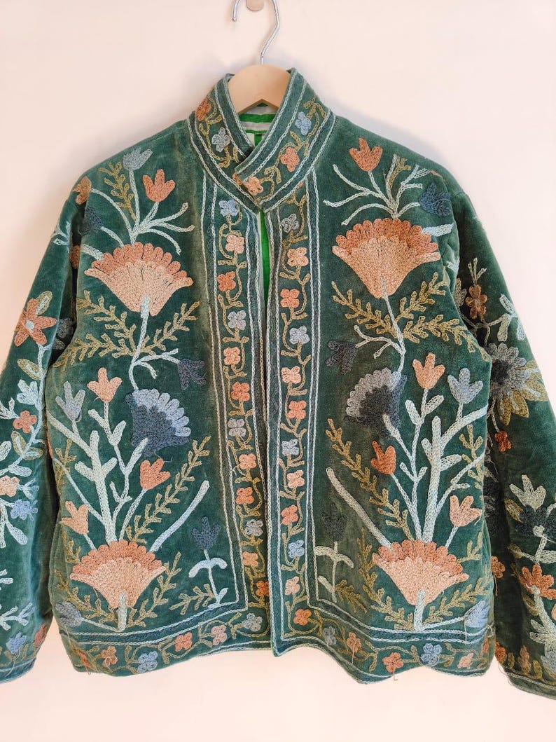 Puede incluir: Chaqueta de terciopelo verde con intrincados bordados florales en tonos melocot&oacute;n, azul y crema. La chaqueta presenta un cuello alto y un dise&ntilde;o bordado detallado en la parte delantera y las mangas. Colgada en una percha de madera.