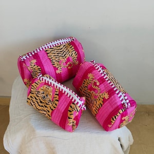 Può includere: Tre trousse per cosmetici rosa e arancioni con stampa di tigre e cerniere bianche. Le borse sono cilindriche e hanno un motivo a righe. Le borse sono esposte su un panno bianco.