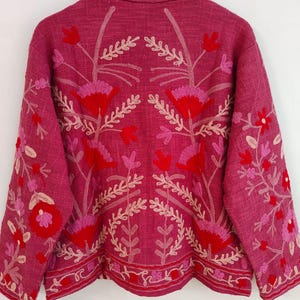 May include: A magenta jacket with floral embroidery in red, pink, and cream. The jacket features a collared neckline and long sleeves. The embroidery includes flowers and leaf patterns, creating a detailed and decorative design. The jacket is hanging on a wooden hanger.
