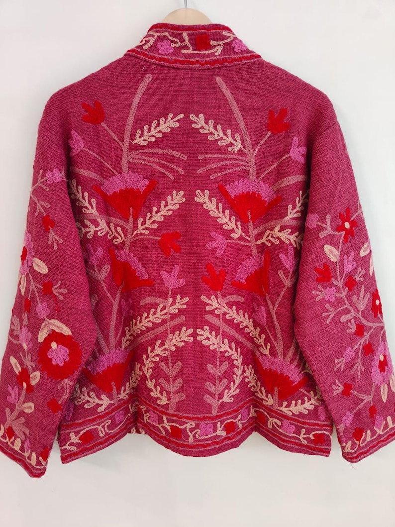 May include: A magenta jacket with floral embroidery in red, pink, and cream. The jacket features a collared neckline and long sleeves. The embroidery includes flowers and leaf patterns, creating a detailed and decorative design. The jacket is hanging on a wooden hanger.