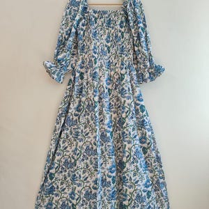 Può includere: Un lungo vestito bianco con motivo floreale blu e maniche lunghe. L'abito presenta uno scollo quadrato, un corpetto aderente e una gonna fluida. Le maniche hanno polsini a volant. L'abito è appeso a una gruccia di legno.