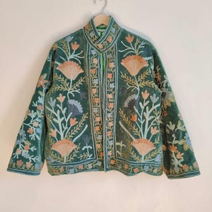 Puede incluir: Chaqueta de terciopelo verde con intrincados bordados florales en tonos melocot&oacute;n, azul y blanco. La chaqueta presenta un cuello mandar&iacute;n y mangas largas. El bordado se concentra en el frente, las mangas y el cuello.