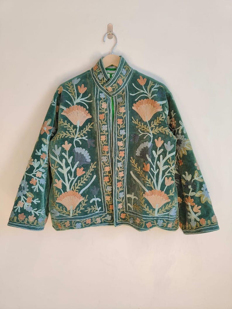 Puede incluir: Chaqueta de terciopelo verde con intrincados bordados florales en tonos melocot&oacute;n, azul y blanco. La chaqueta presenta un cuello mandar&iacute;n y mangas largas. El bordado se concentra en el frente, las mangas y el cuello.