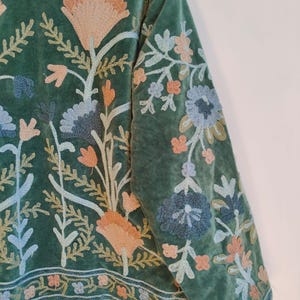 Puede incluir: Chaqueta de terciopelo verde con intrincado bordado floral en tonos melocot&oacute;n, azul y crema. El bordado cubre las mangas y el frente, con un borde detallado en el dobladillo. El dise&ntilde;o de la chaqueta presenta un tema bot&aacute;nico.