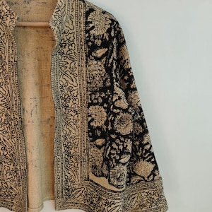 K&ouml;nnte beinhalten: Beige-schwarze Jacke mit floralem Muster. Die Jacke ist offen und zeigt ein beiges Innenfutter. Die &Auml;rmel und Kanten sind mit komplizierten schwarzen Mustern verziert. Die Jacke h&auml;ngt an einem Holzb&uuml;gel.