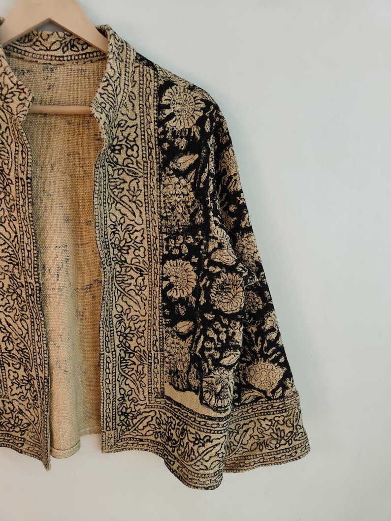 K&ouml;nnte beinhalten: Beige-schwarze Jacke mit floralem Muster. Die Jacke ist offen und zeigt ein beiges Innenfutter. Die &Auml;rmel und Kanten sind mit komplizierten schwarzen Mustern verziert. Die Jacke h&auml;ngt an einem Holzb&uuml;gel.